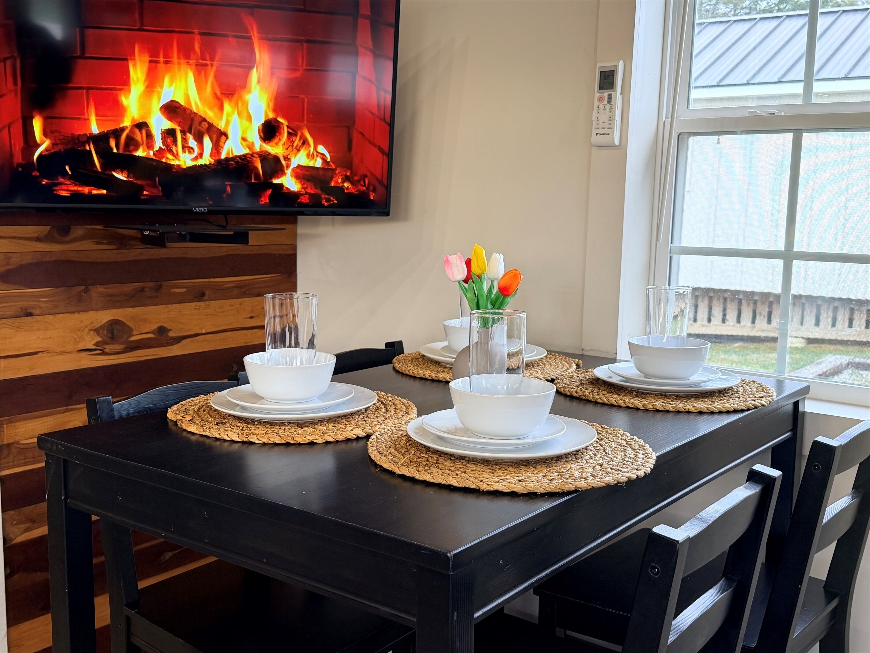 Una mesa de comedor dispuesta con cuencos y platos blancos, una copa y un jarrón con tulipanes. Se ve una chimenea con un televisor al fondo.