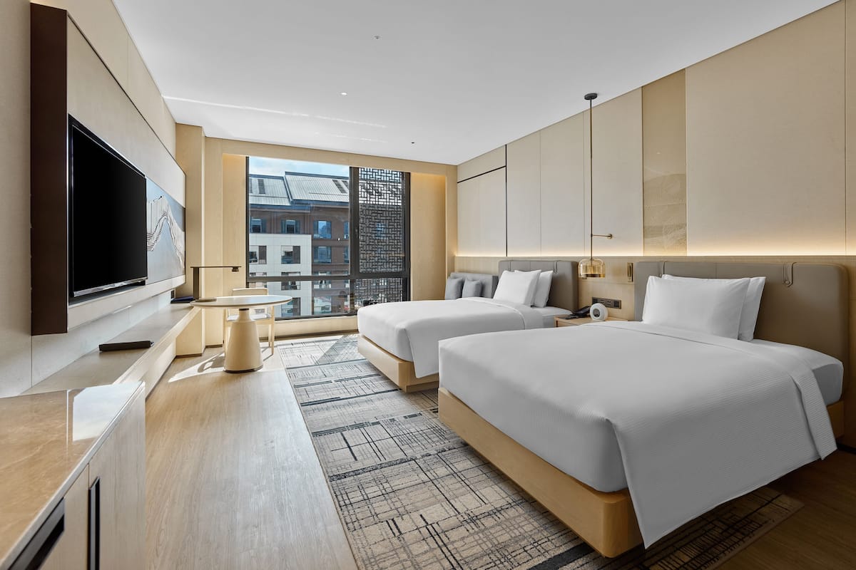 A modern hotel room with two beds, a flat-screen TV, a desk, and a large window offering a city view.
