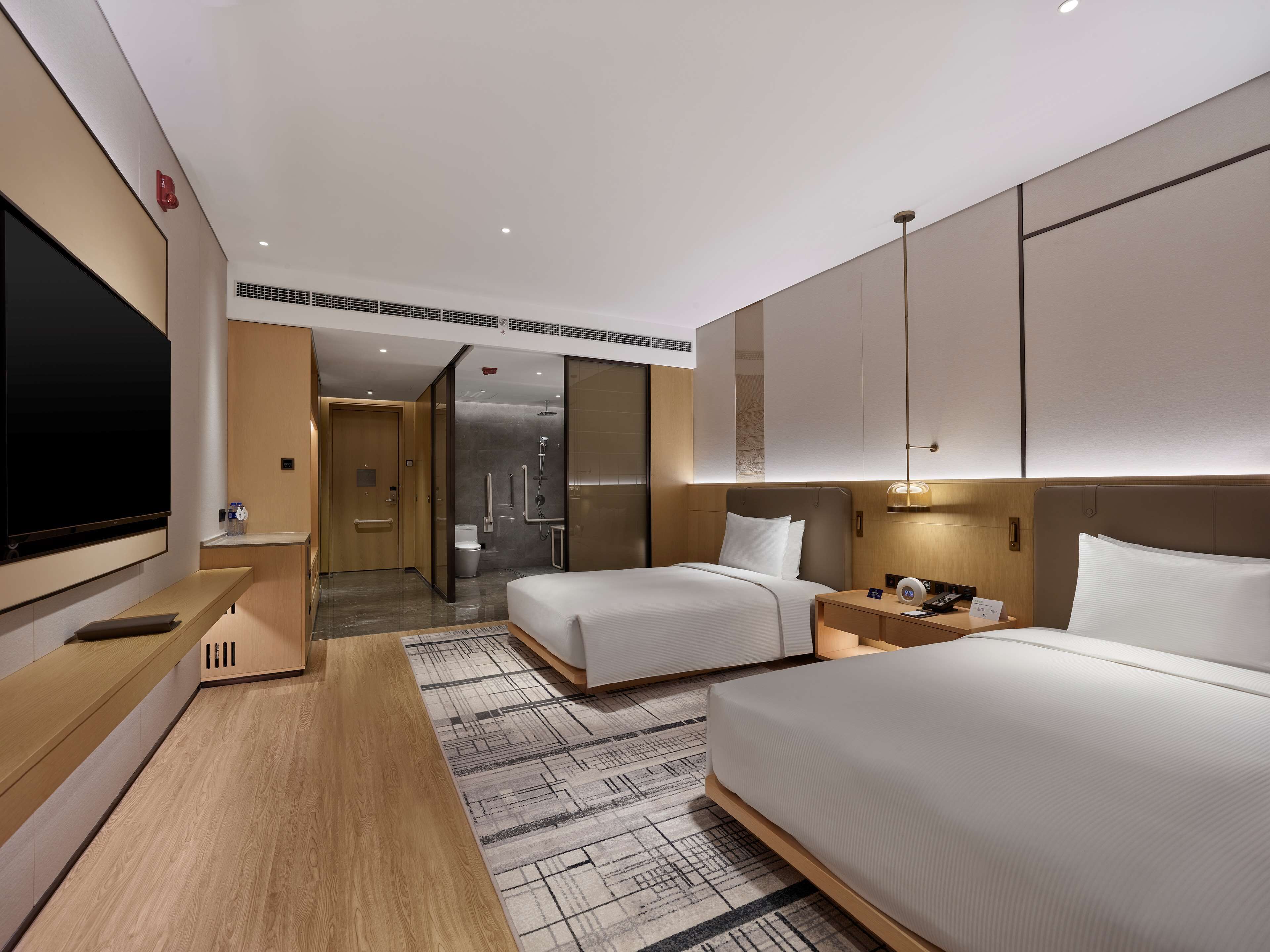 Une chambre d’hôtel moderne équipée de deux lits, d’un téléviseur à écran plat, d’une table de chevet et d’une salle de bain avec douche et toilettes.
