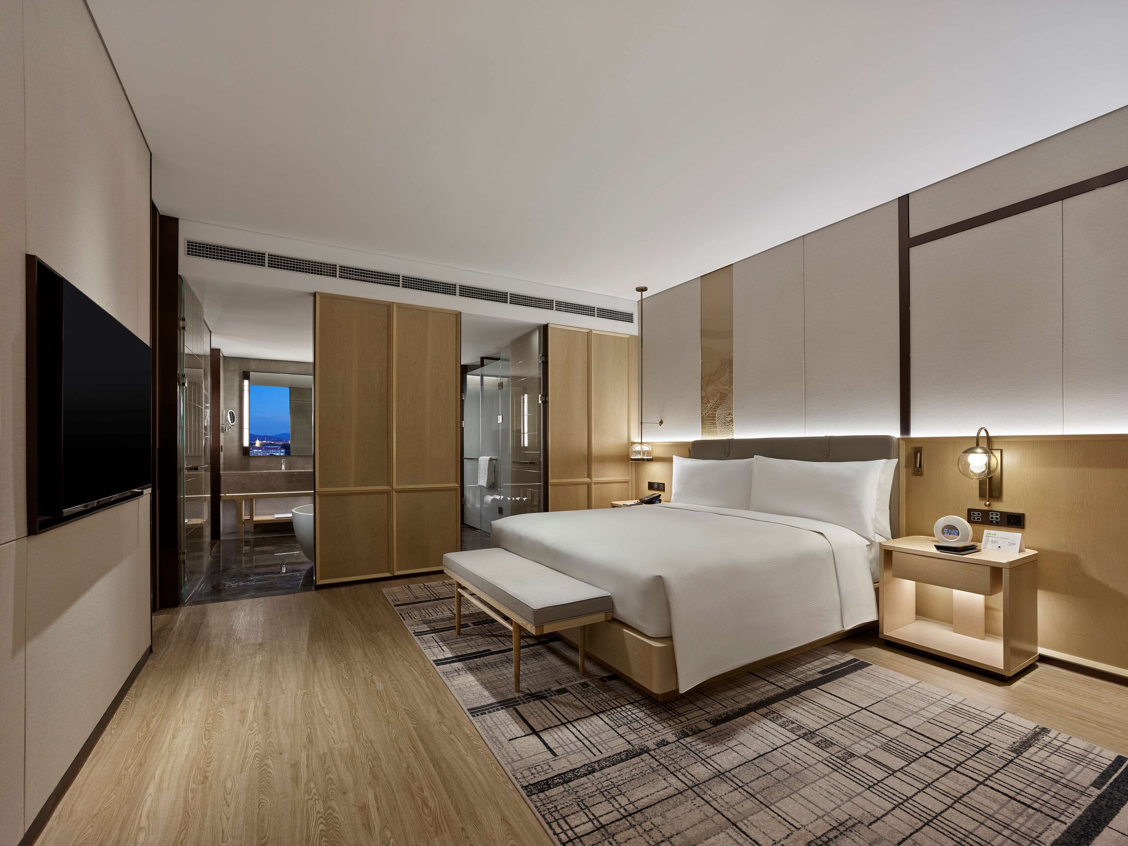 A modern hotel room with a large bed, bedside tables, a TV, and a view of the city.