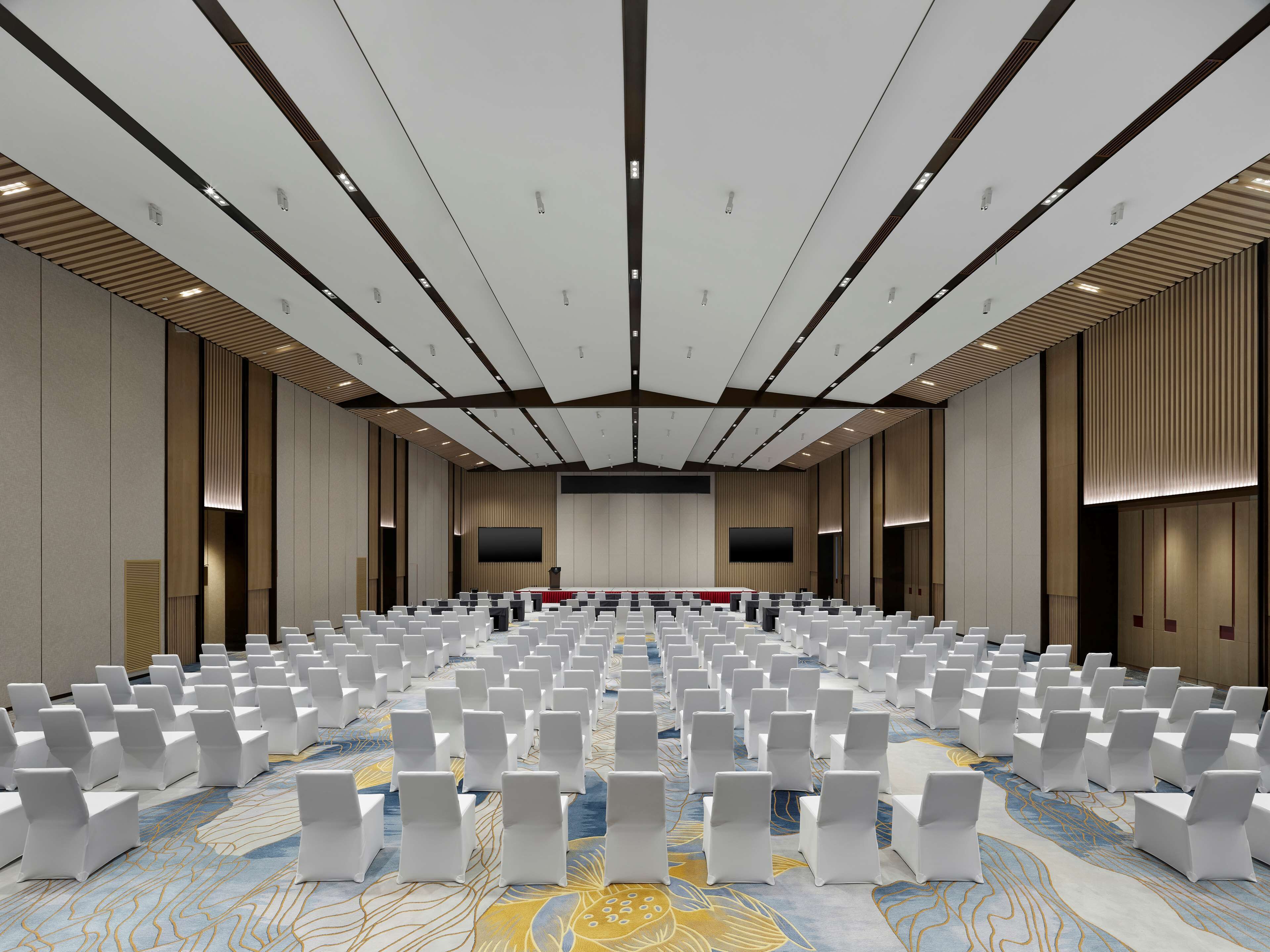 Un salón de conferencias amplio con filas de sillas blancas y un escenario al frente.