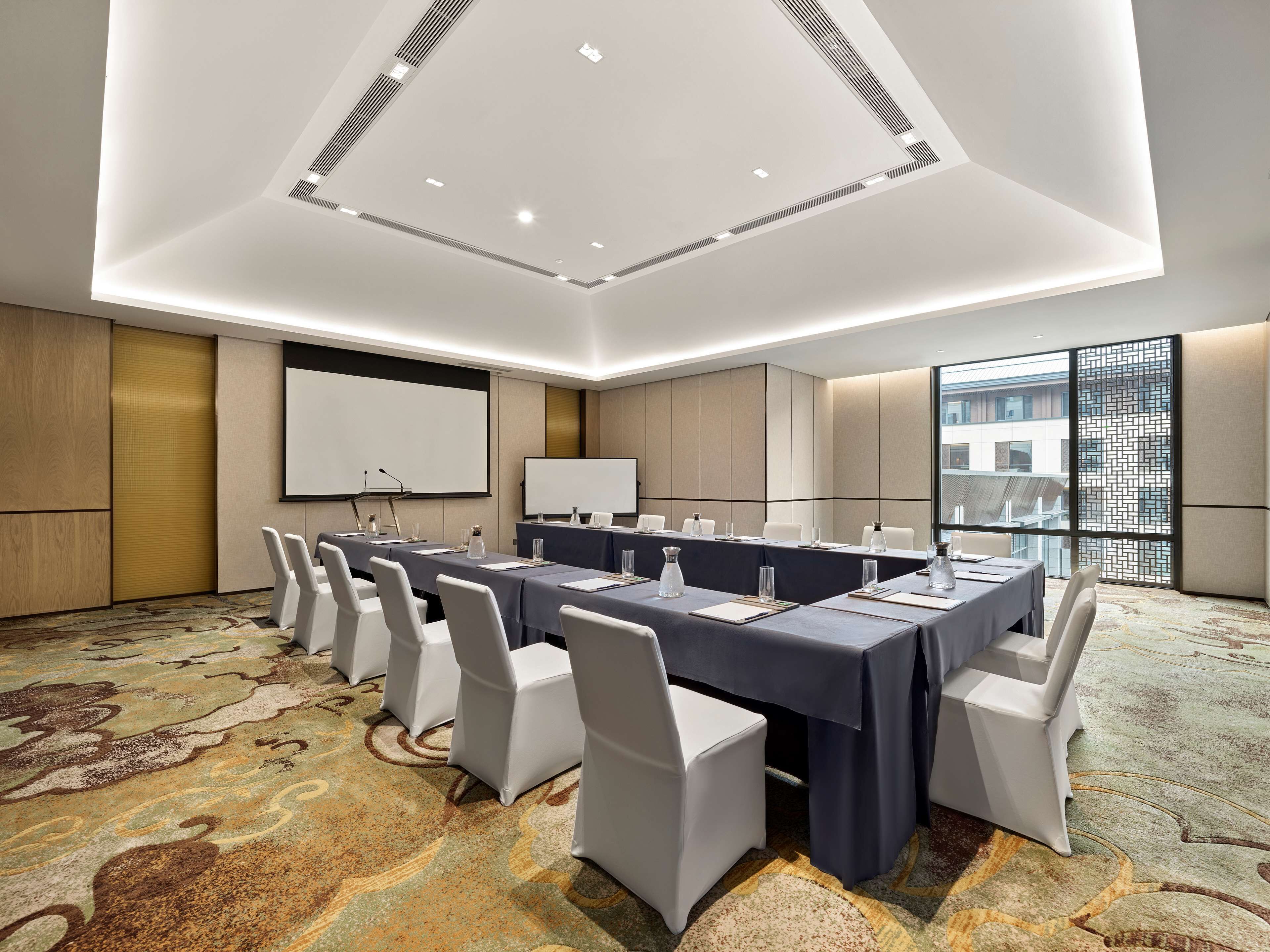 Une salle de conférence avec une longue table, des chaises blanches, un écran de projection et une grande fenêtre donnant sur des immeubles.
