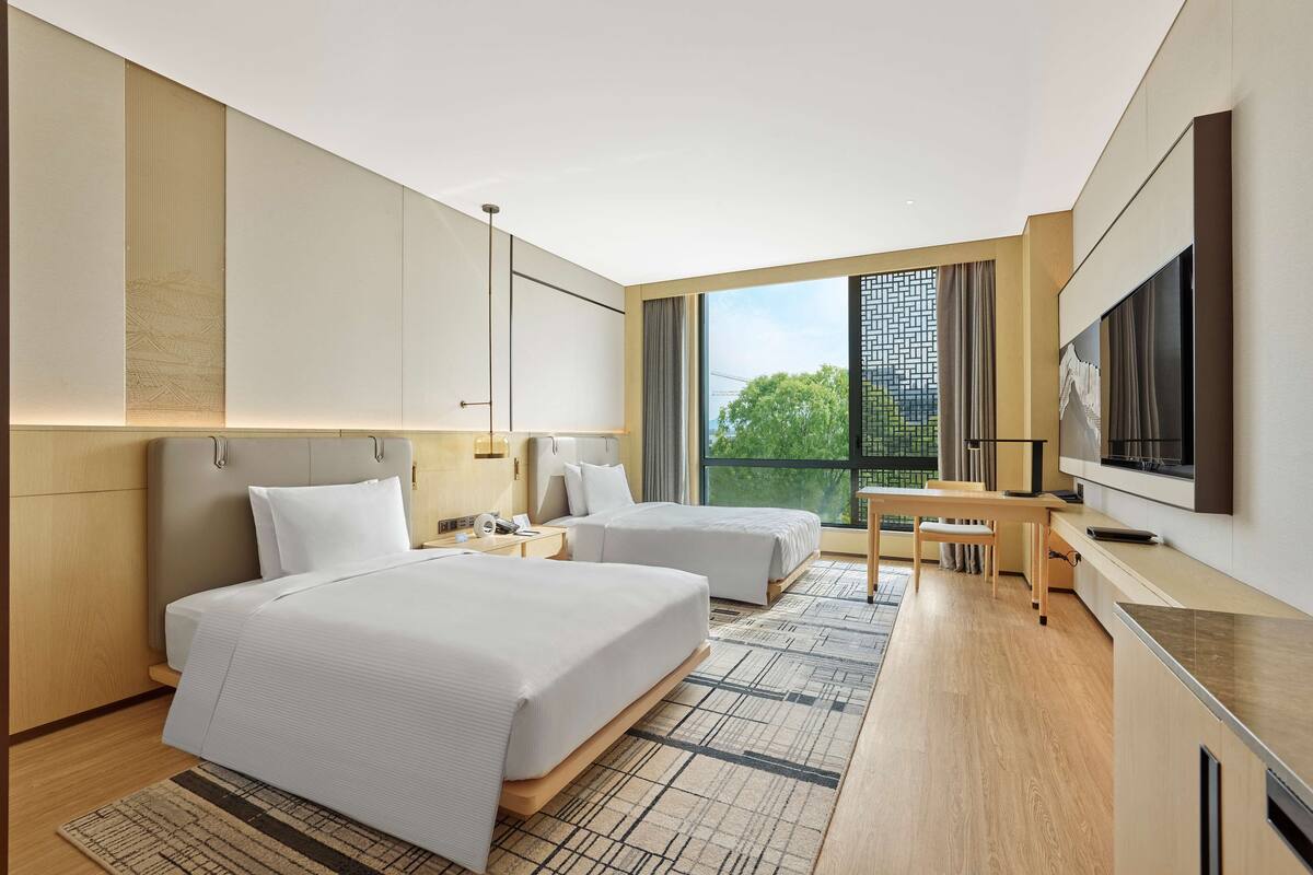 A modern hotel room with two beds, a desk, and a large window offering a view of greenery.