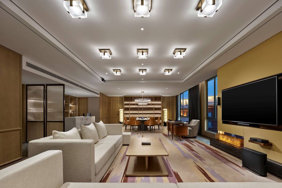 A modern hotel room with a sofa, a coffee table, a TV, and a dining area with chairs and a table.
