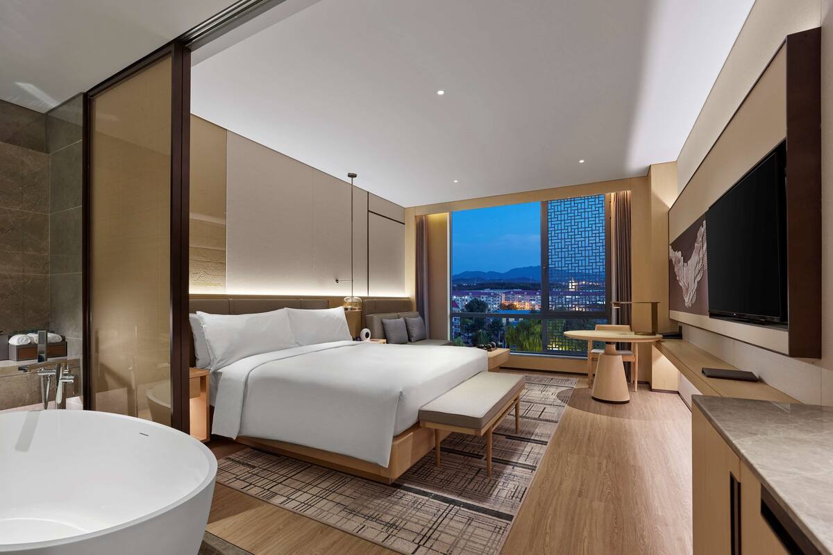 A modern hotel room with a large bed, a bathtub, a desk, and a view of the cityscape.