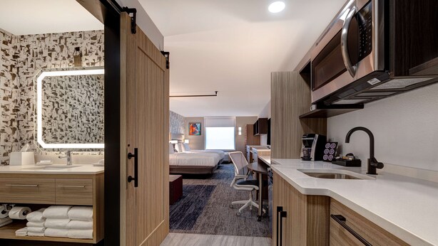 A modern hotel room with a kitchenette, a large mirror, a bed, and a desk area.