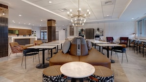 A modern hotel lobby with a central seating area, multiple tables, and a reception desk.