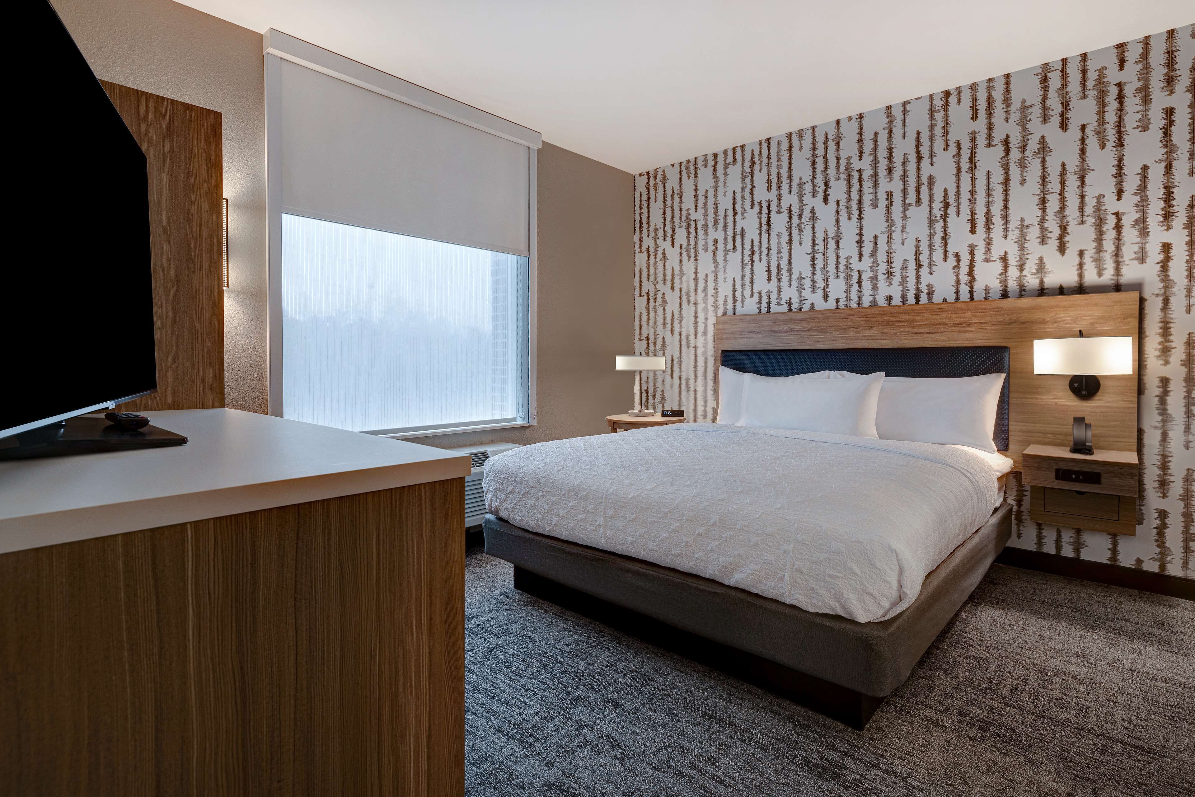 Ein Hotelzimmer mit einem großen Bett, einem Schreibtisch, einem Fernseher und einem Fenster mit Jalousien.