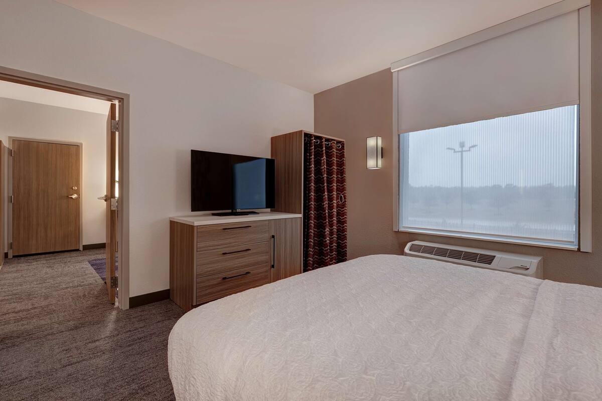 Une chambre d’hôtel avec un lit, un téléviseur posé sur une commode en bois, une fenêtre avec des stores et une porte donnant sur une autre pièce.