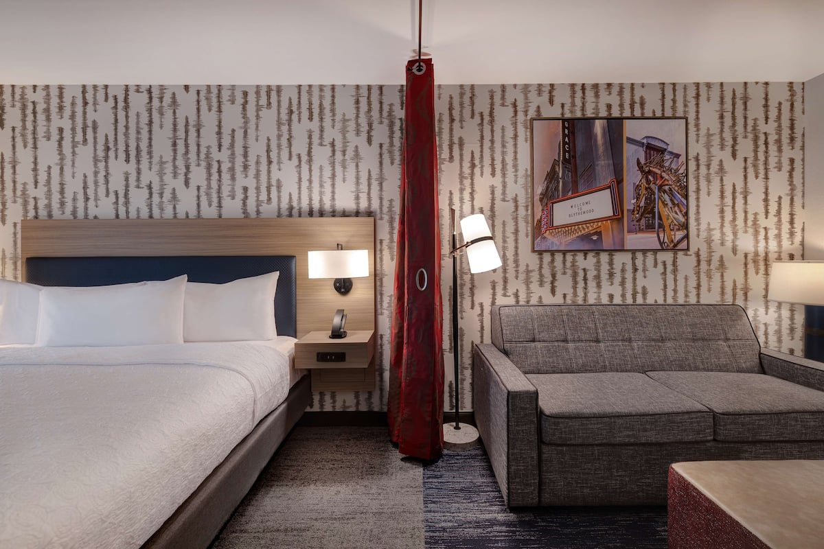 Une chambre d’hôtel avec un lit, une table de chevet avec une lampe, un canapé et un mur orné d’un motif de forêt ainsi qu’une pancarte de bienvenue.