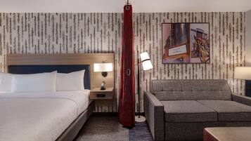 A hotel room with a bed, a nightstand with a lamp, a sofa, and a wall with a forest pattern and a welcome sign.
