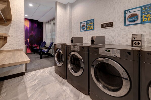 A modern laundry facility with two front-loading washers and dryers, a wooden counter, and a wall-mounted sign indicating the location of the front desk.