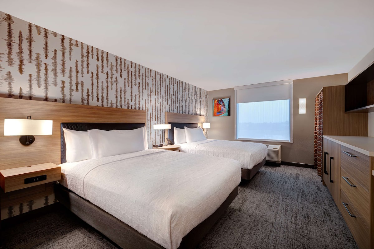 Une chambre d’hôtel avec deux lits, une grande fenêtre, un téléviseur et un mur décoré d’un motif représentant un arbre.