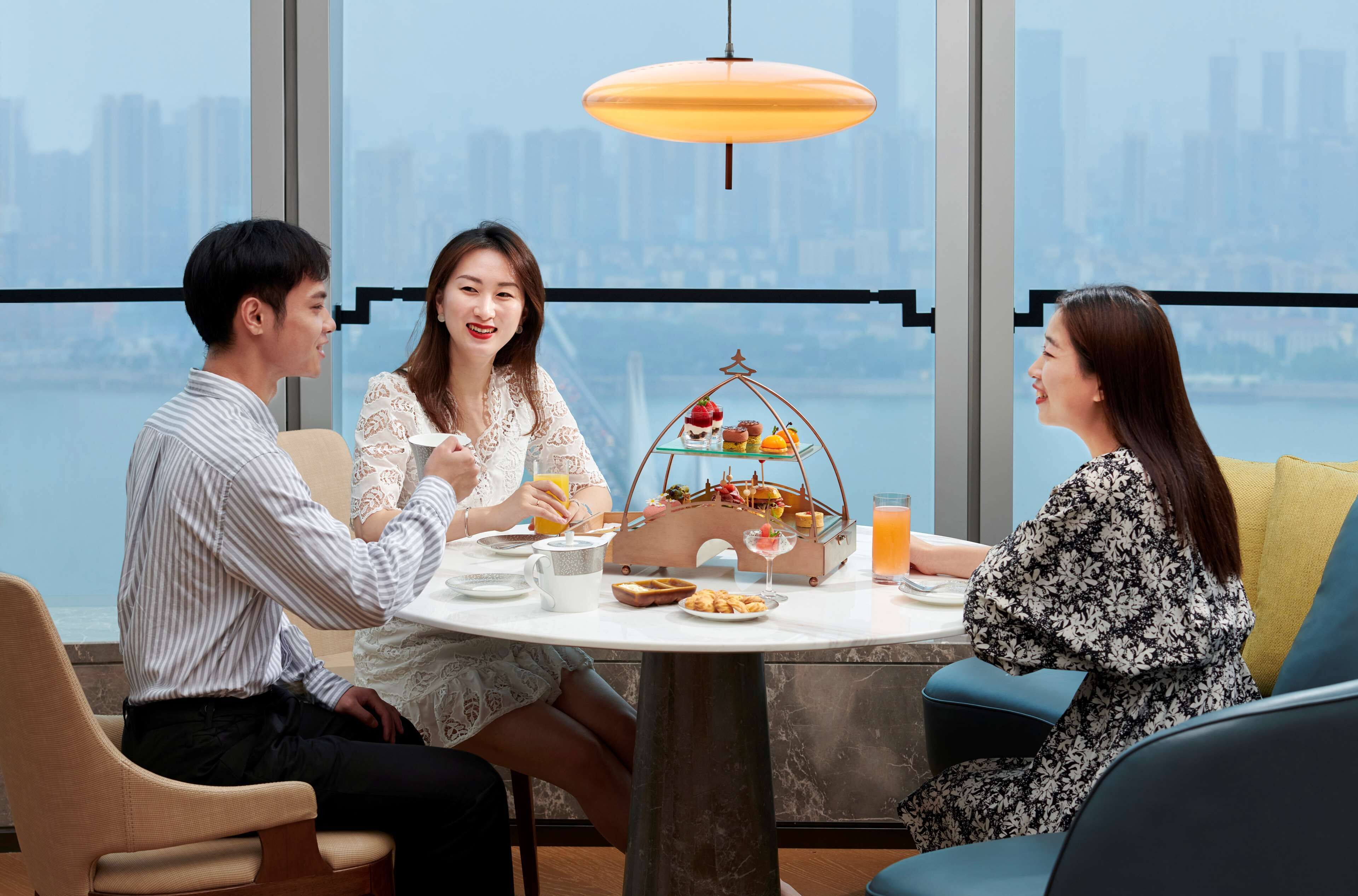 A dining area with a table set for three, a fruit display, and a city view through large windows.