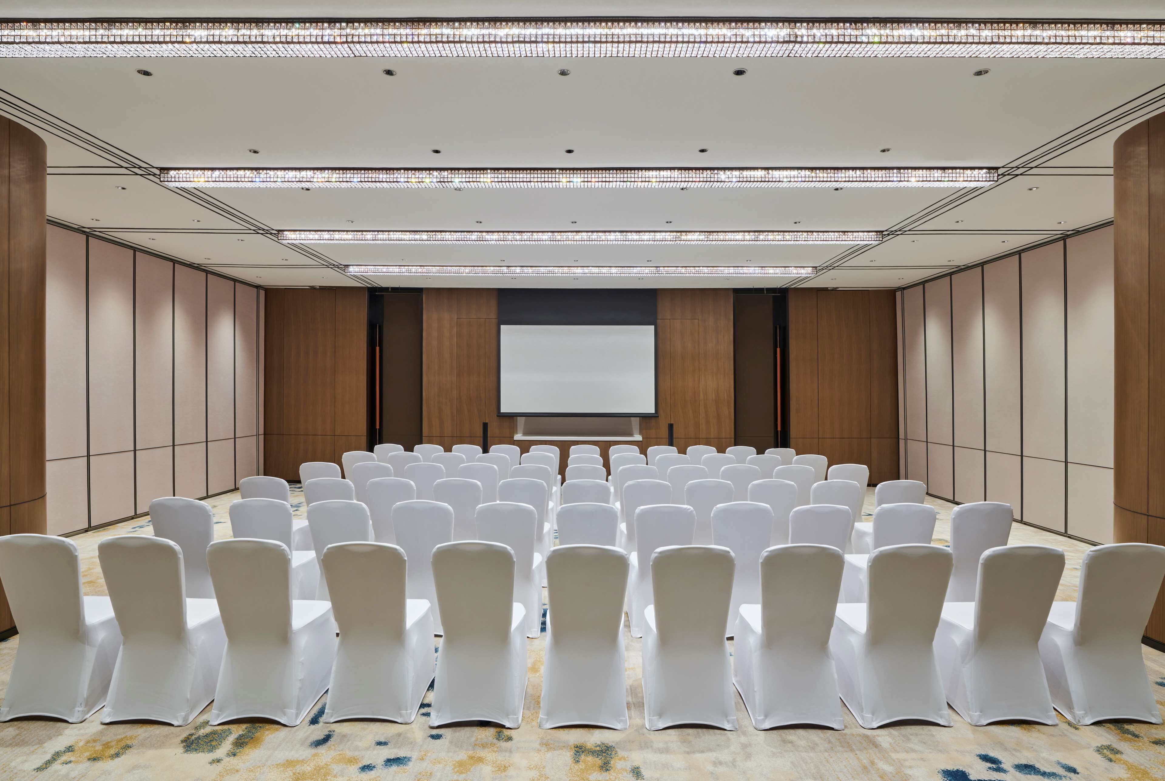 Una sala conferenze con file di sedie bianche, uno schermo per proiettore e un pavimento rivestito di moquette.