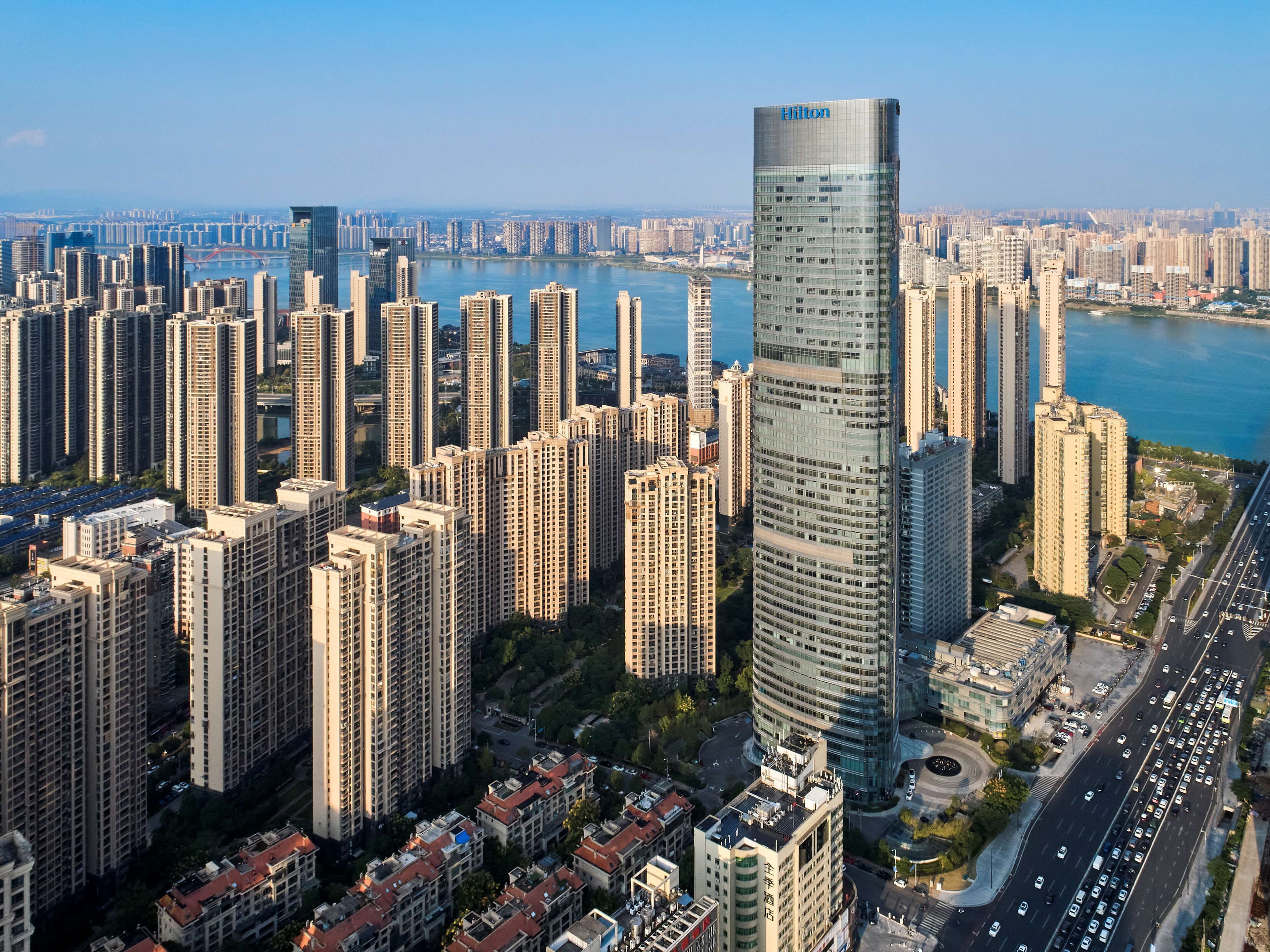 Un hotel moderno de gran altura con fachada de vidrio, rodeado de edificios urbanos y un río al fondo.