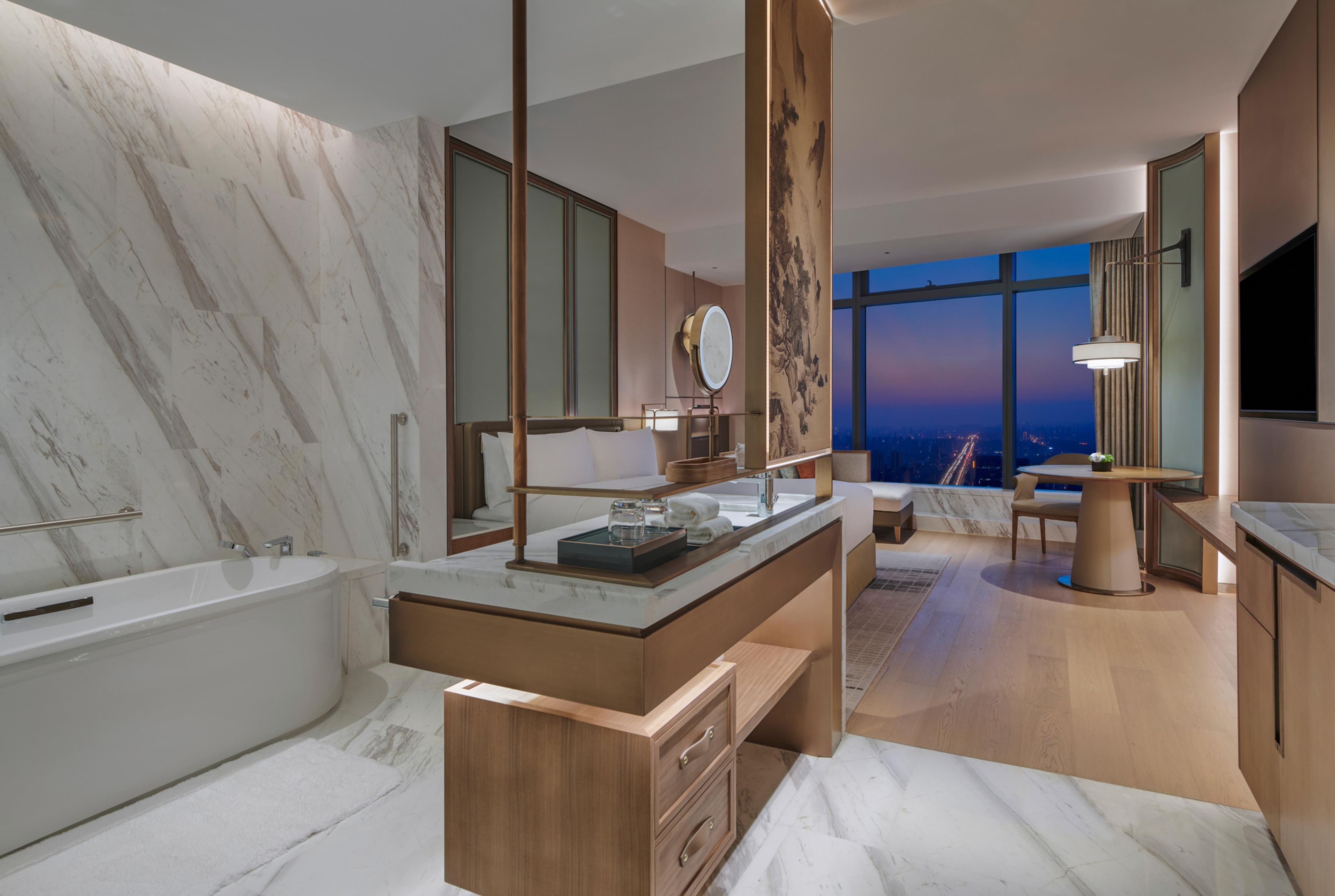 Un baño moderno con pared de mármol, una bañera, un tocador con espejo y vistas a la ciudad al anochecer.