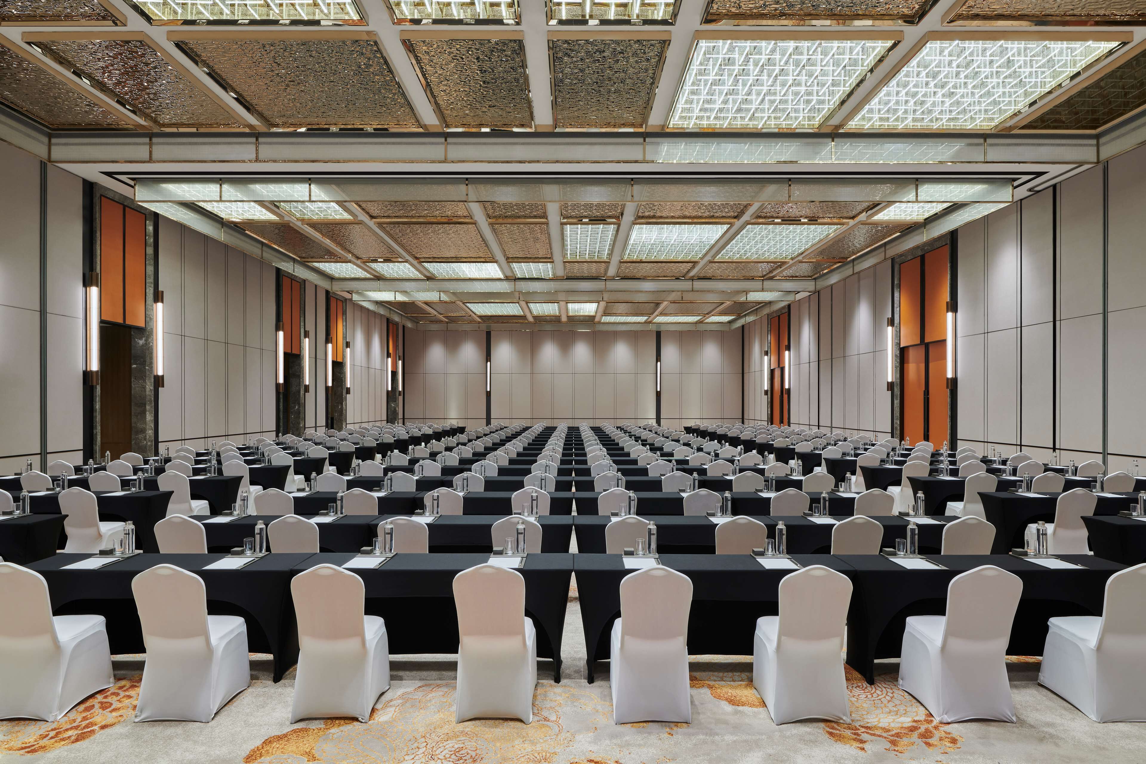 Een congreszaal met rijen tafels en stoelen, grote ramen en een hoog plafond met een rasterpatroon.