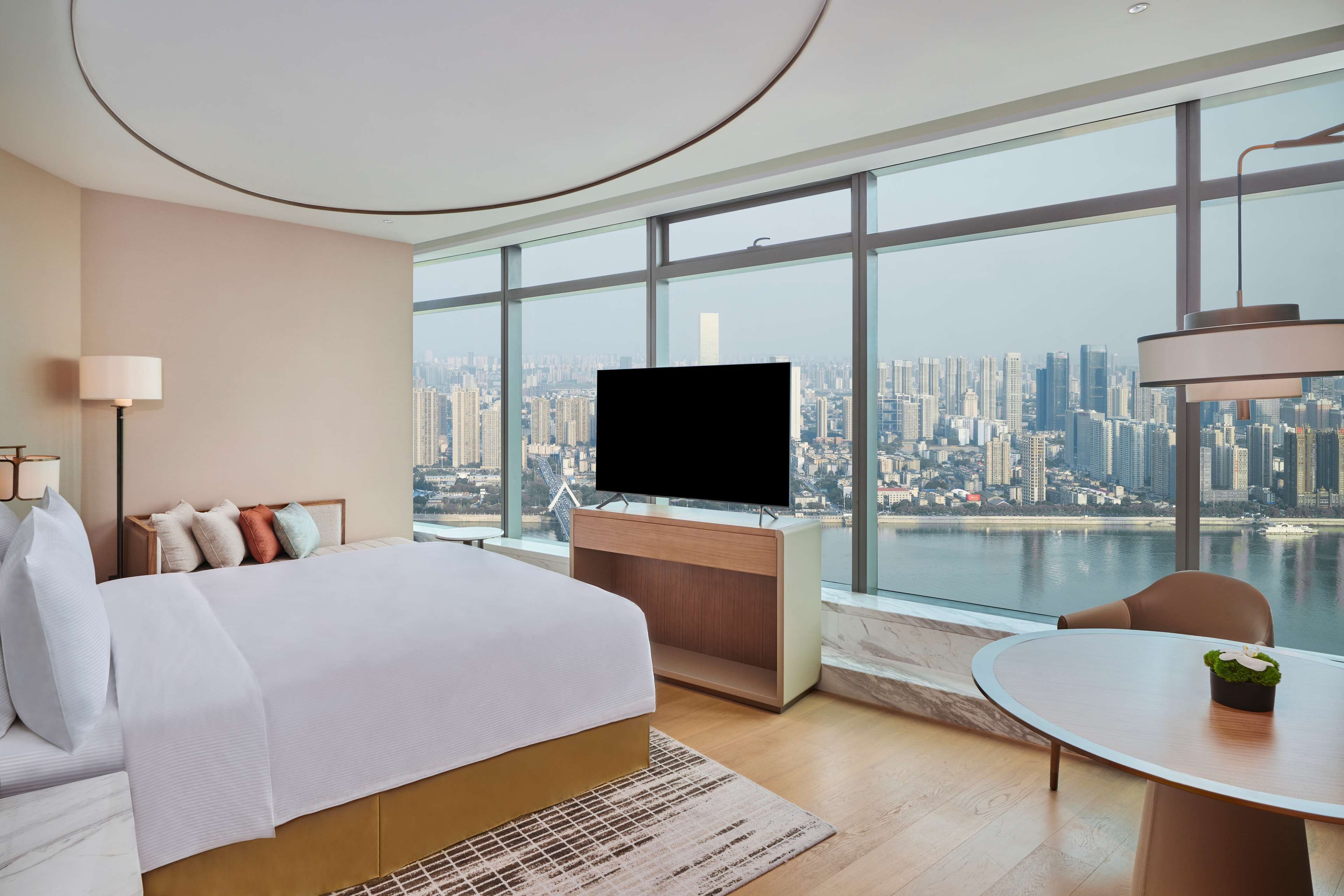 A modern hotel room with a large bed, a desk with a flat-screen TV, a round table with a plant, and a city view through large windows.