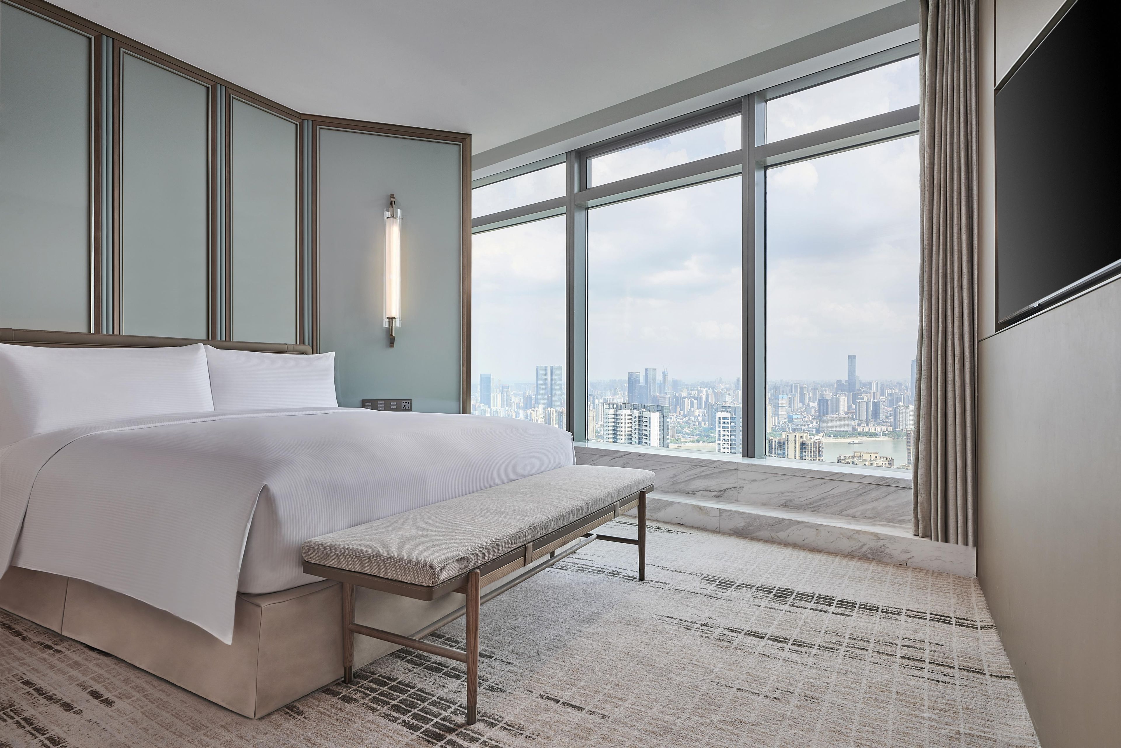 A modern hotel room with a large bed, a bench, a TV, and a city view.