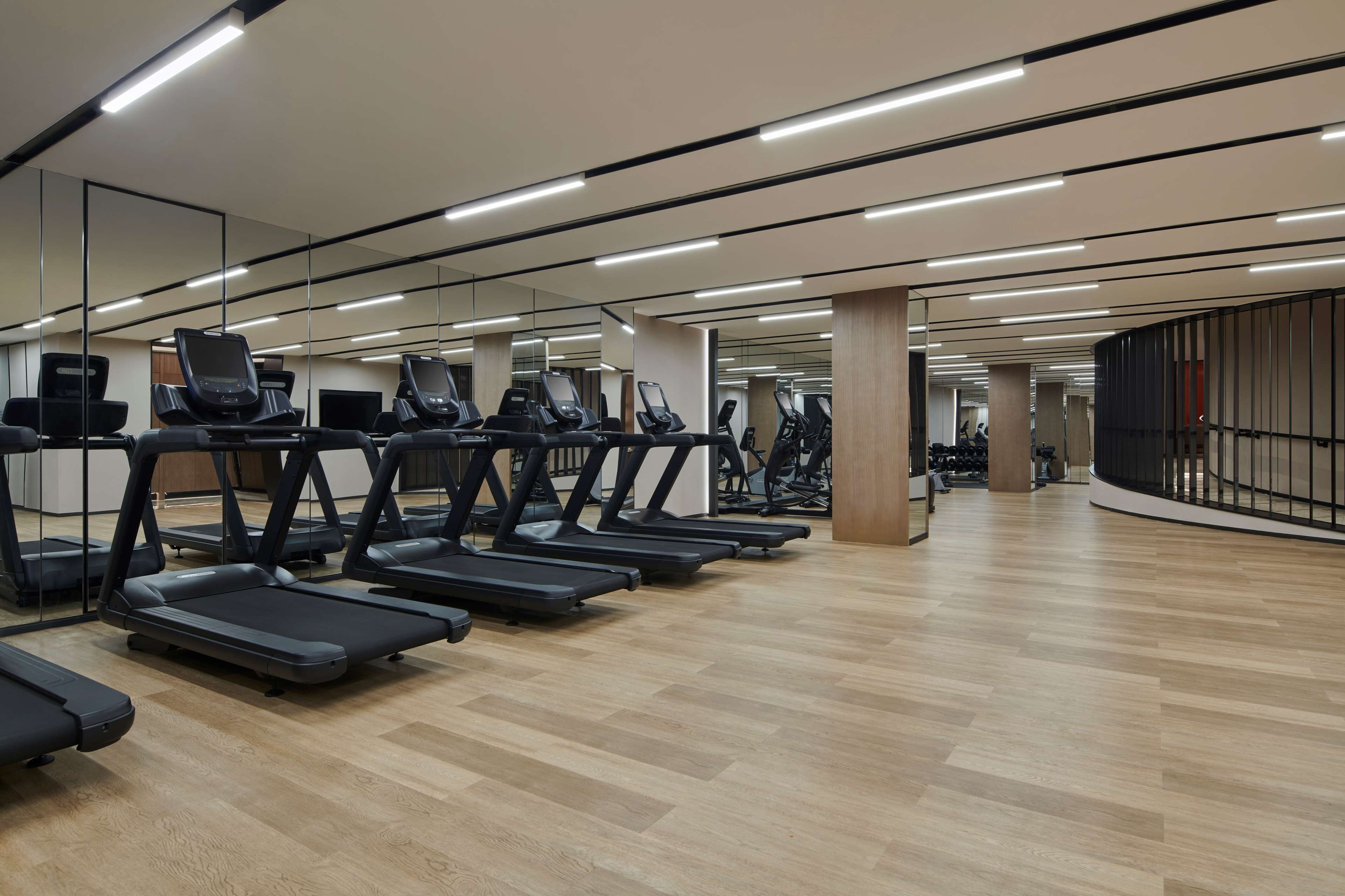 A gym with multiple treadmills lined up against a mirrored wall, wooden flooring, and a curved wall with a railing.