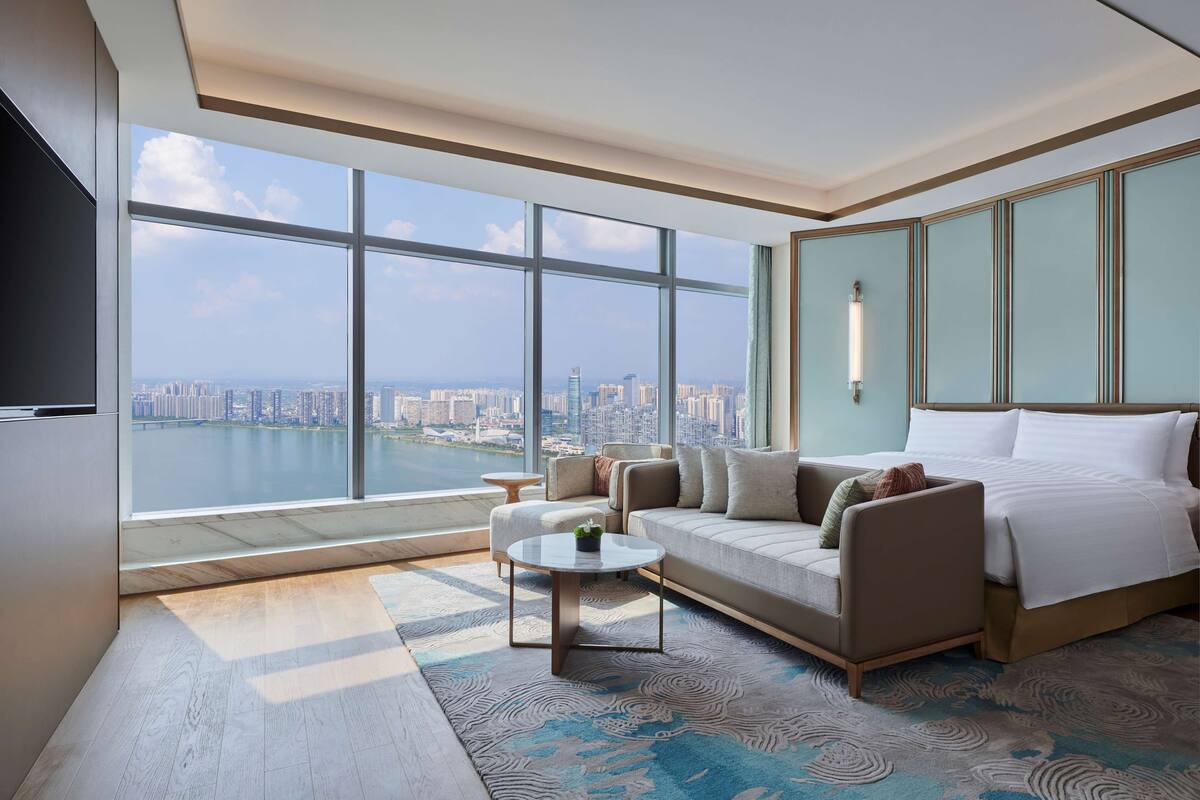 A modern hotel room with a large window offering a city view, a sofa, a bed, and a small table.