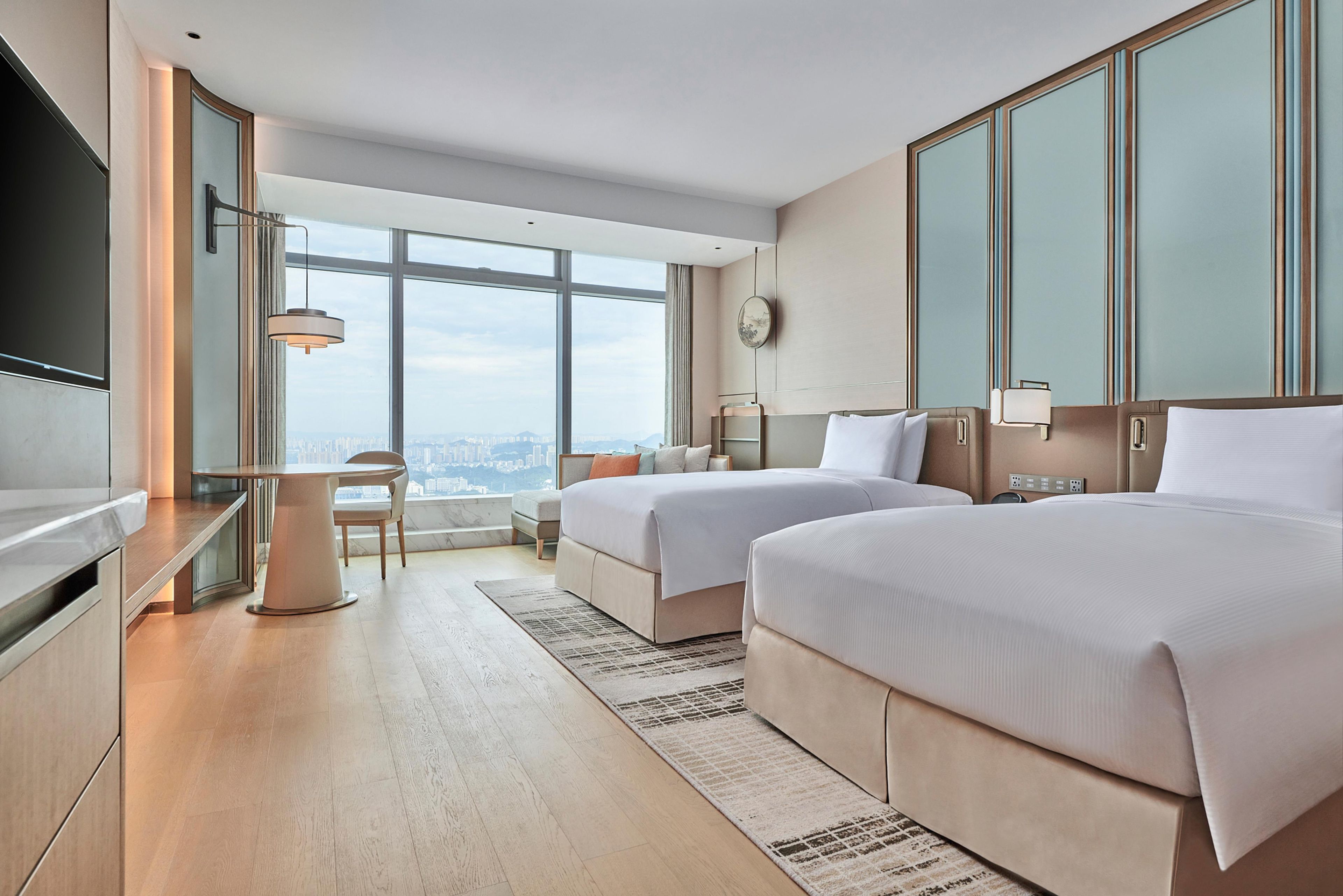 A modern hotel room with two beds, a desk, a chair, and a large window offering a city view.