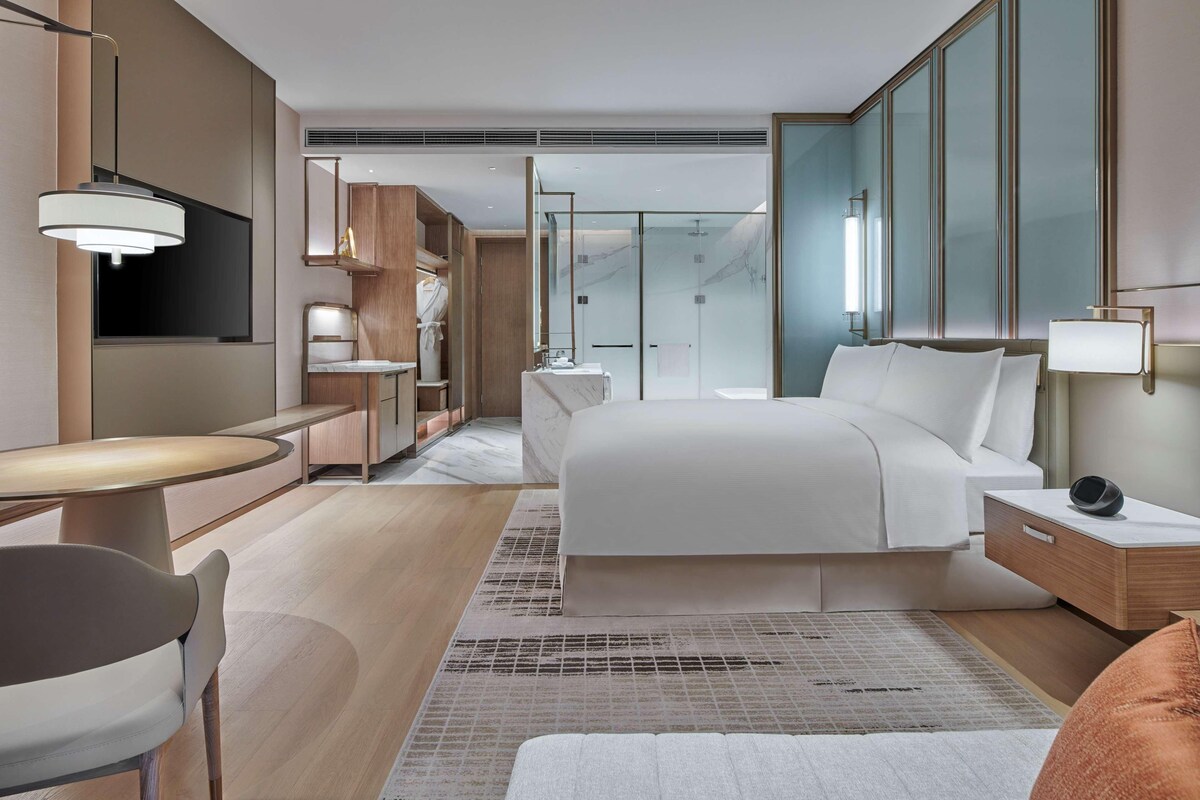 A modern hotel room with a bed, bedside table, lamp, and a seating area with a chair and table.