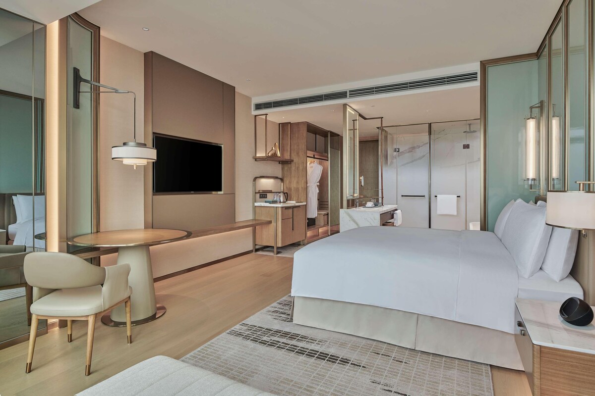A modern hotel room with a large bed, a desk, a chair, a TV, and a walk-in closet.