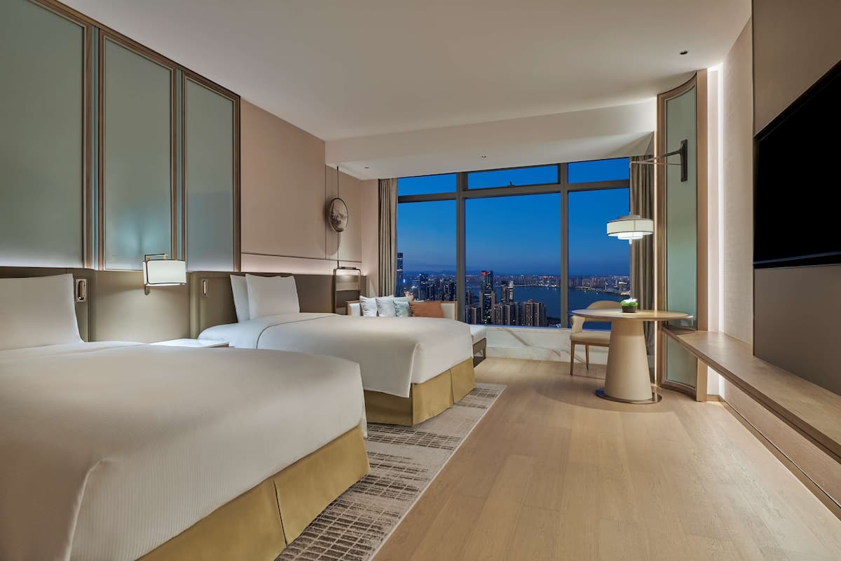 A hotel room with two beds, a desk, a chair, a TV, and a large window with a city view.