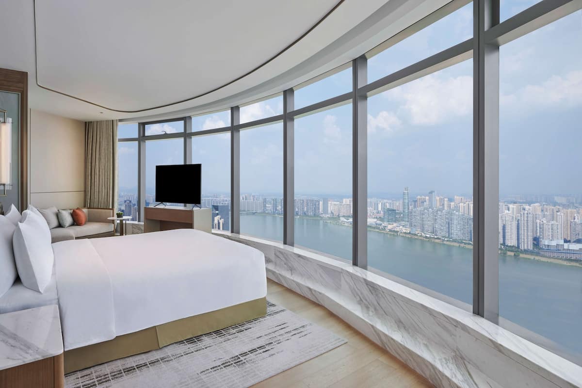 A modern hotel room with a curved ceiling, a large bed, a flat-screen TV, and a view of a cityscape and river.