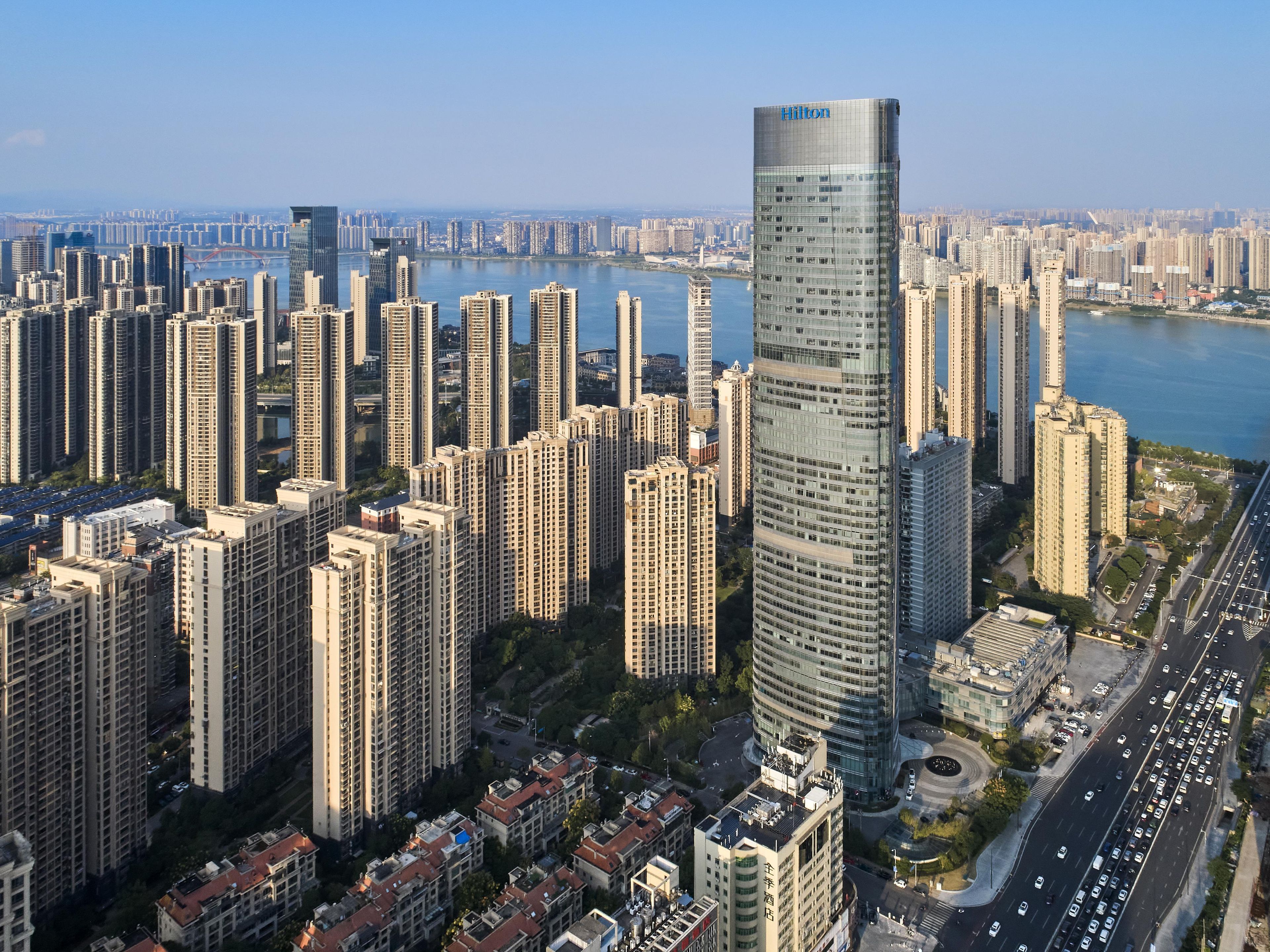 A high-rise hotel with a modern glass facade, surrounded by dense urban high-rise buildings, and a busy road with vehicles.