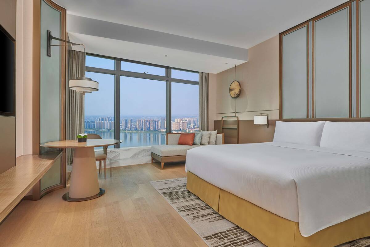 A modern hotel room with a large bed, a desk, and a view of the city through large windows.