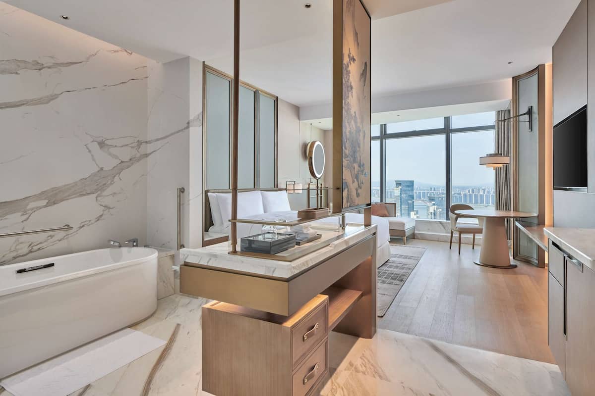 A modern bathroom with a marble wall, a bathtub, a vanity with a mirror, and a city view through large windows.