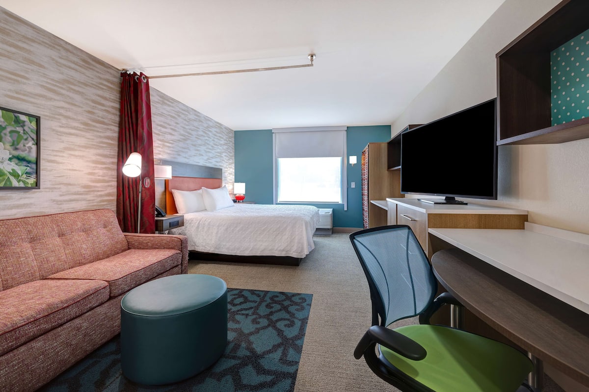 A hotel room with a bed, a sofa, a desk with a chair, a TV, and a window with curtains.