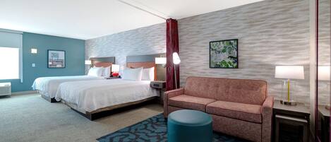 A hotel room with two beds, a sofa, a round ottoman, and a nightstand with a lamp.
