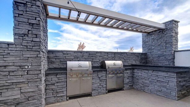 Une terrasse extérieure dotée de deux barbecues en acier inoxydable, d’un mur en pierre et d’une structure en forme de pergola.