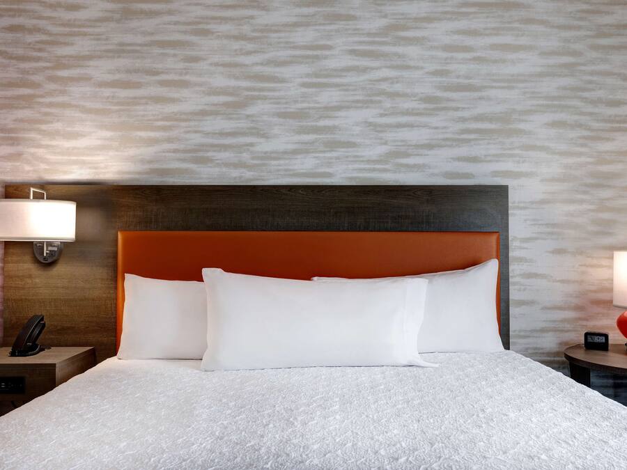 A hotel room with a large bed, two bedside tables, a nightstand, a red lamp, and a wall-mounted lamp.