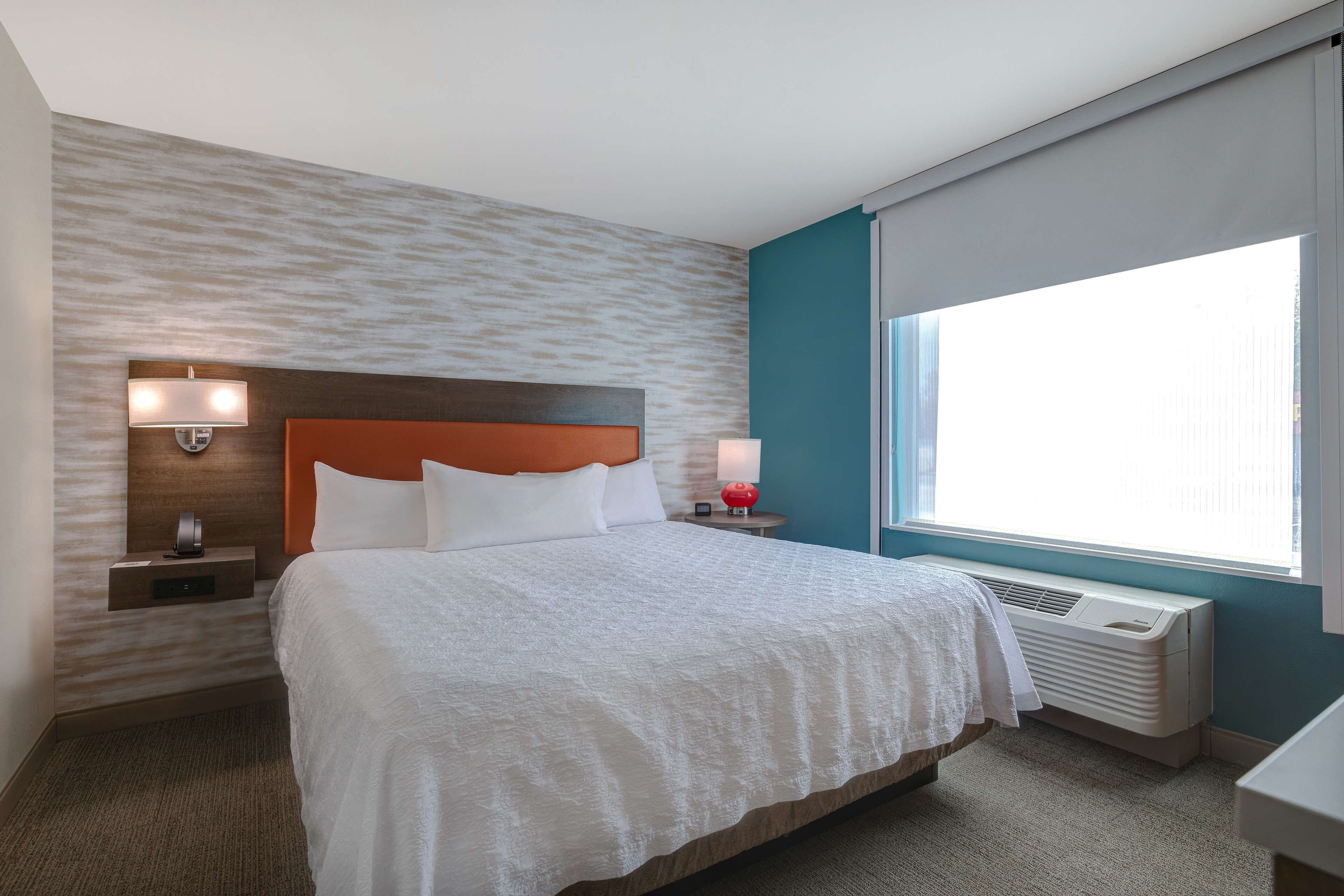 A hotel room with a bed, nightstands, a wall-mounted heater, and a window with curtains.