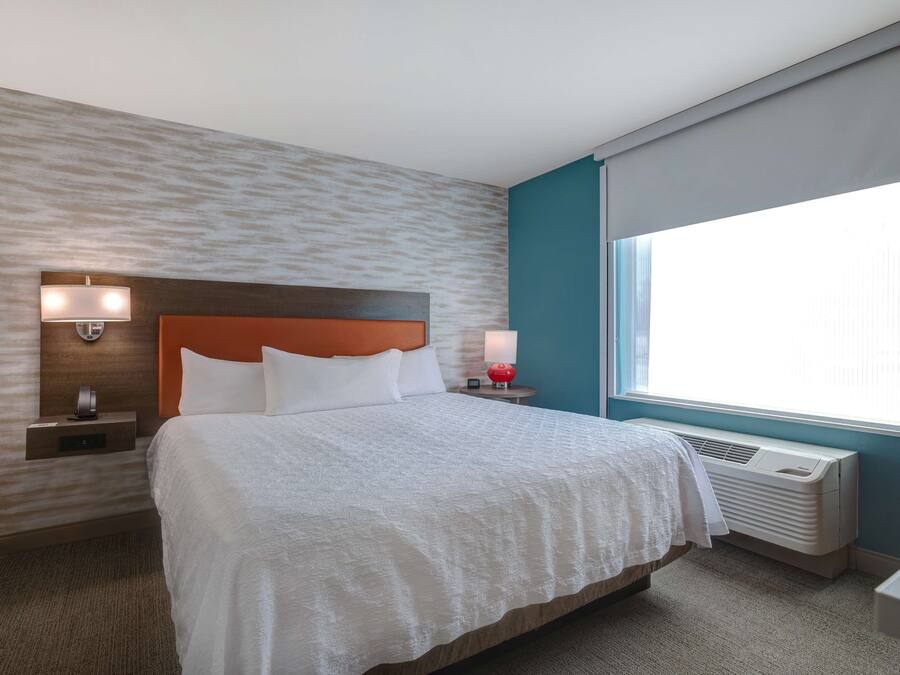 A hotel room with a bed, nightstands, a wall-mounted heater, and a window with curtains.