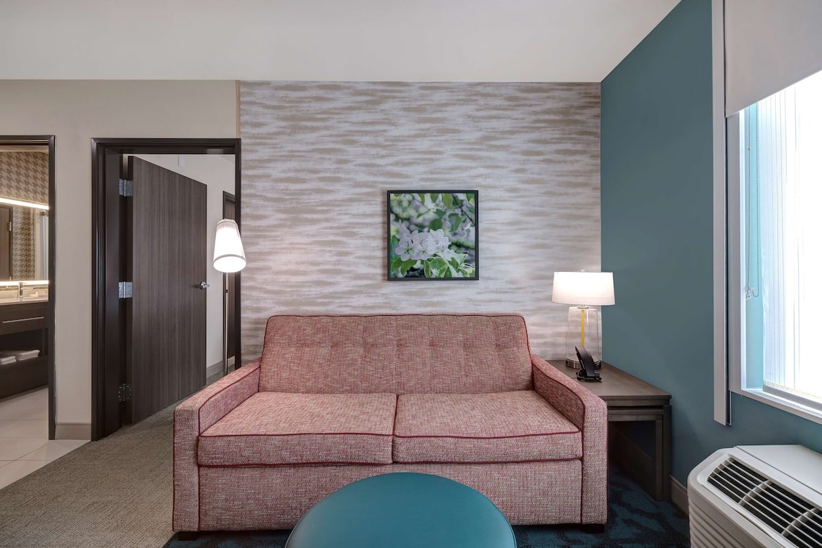 A hotel room with a red sofa, two bedside lamps, a framed picture on the wall, a window with blinds, and a wall-mounted air conditioner.