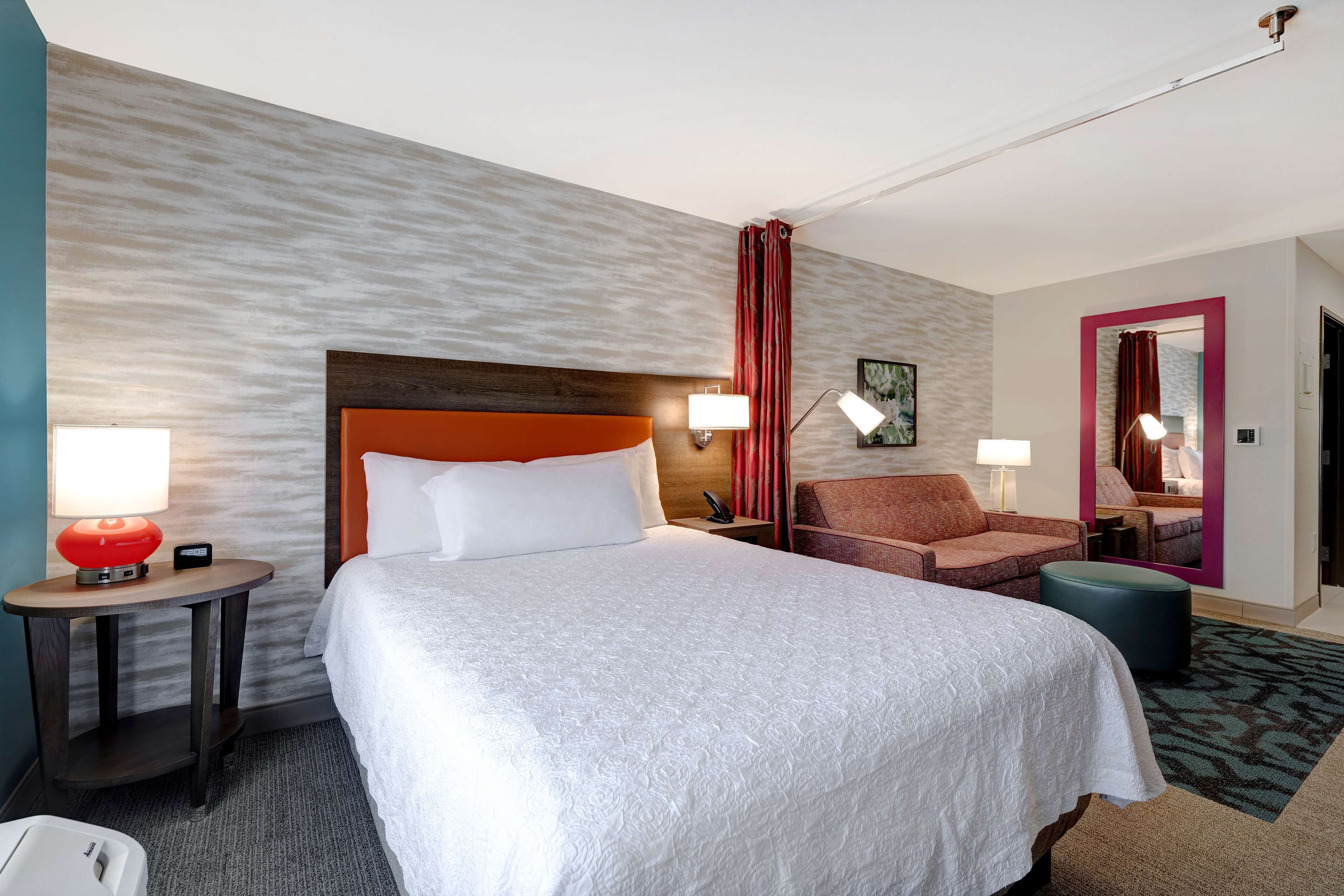 A hotel room with a large bed, a wooden headboard, two bedside tables with lamps, a red chair, a round ottoman, and a mirror with a pink frame.