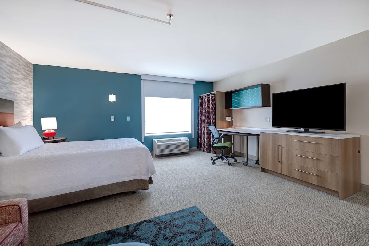 A hotel room with a bed, desk, TV, and a window with curtains.