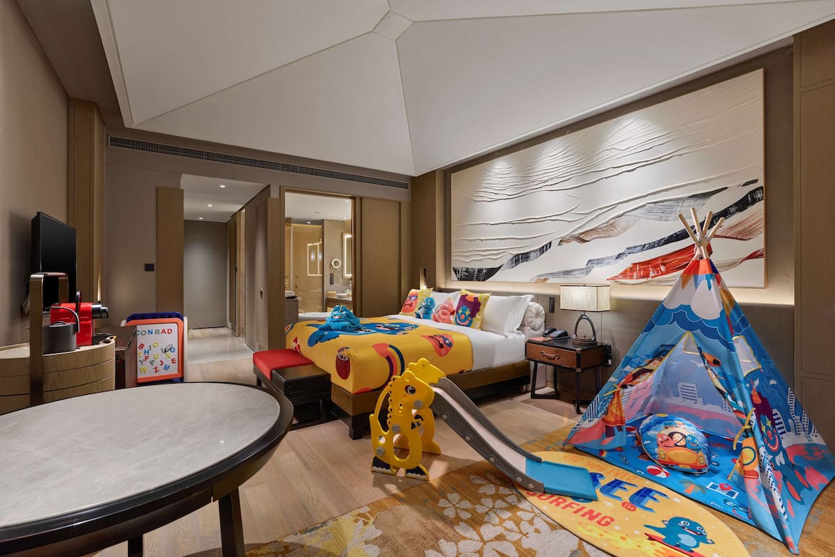 A hotel room with a bed, a desk, a chair, a table, a sliding toy, a tent, and a wall with a painting.
