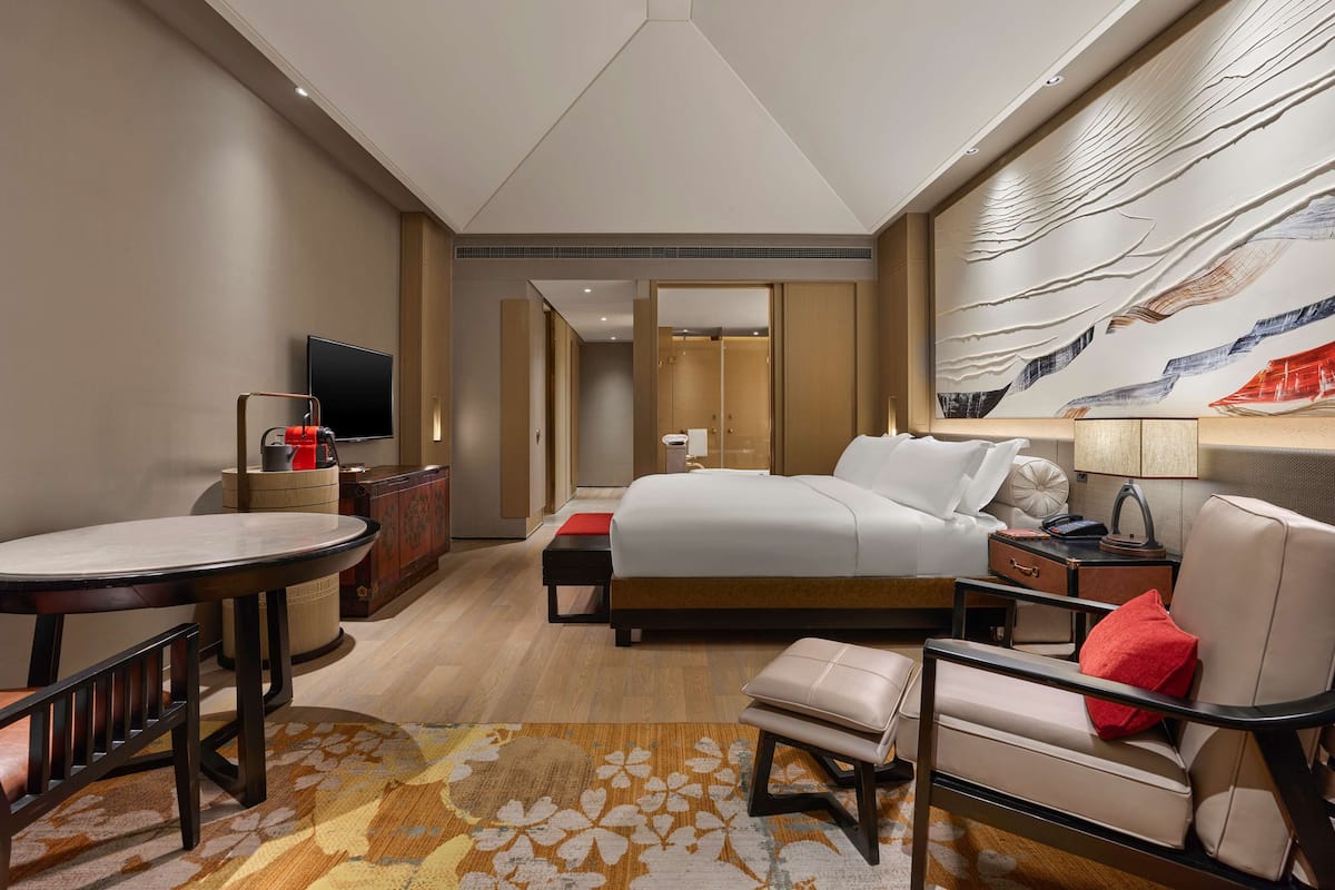 A modern hotel room with a large bed, a desk with a television, a seating area with a chair and ottoman, and a decorative wall mural.