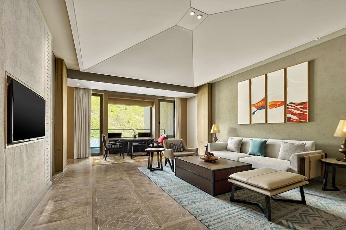 A modern hotel room with a large TV, a sofa, a coffee table, and a dining area with chairs and a table.
