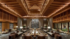 A spacious lobby with a high ceiling and wooden beams, featuring a central seating area with sofas and round tables, flanked by large windows and a stone wall backdrop. - Conrad Jiuzhaigou (Ngawa Autonomous Prefecture)