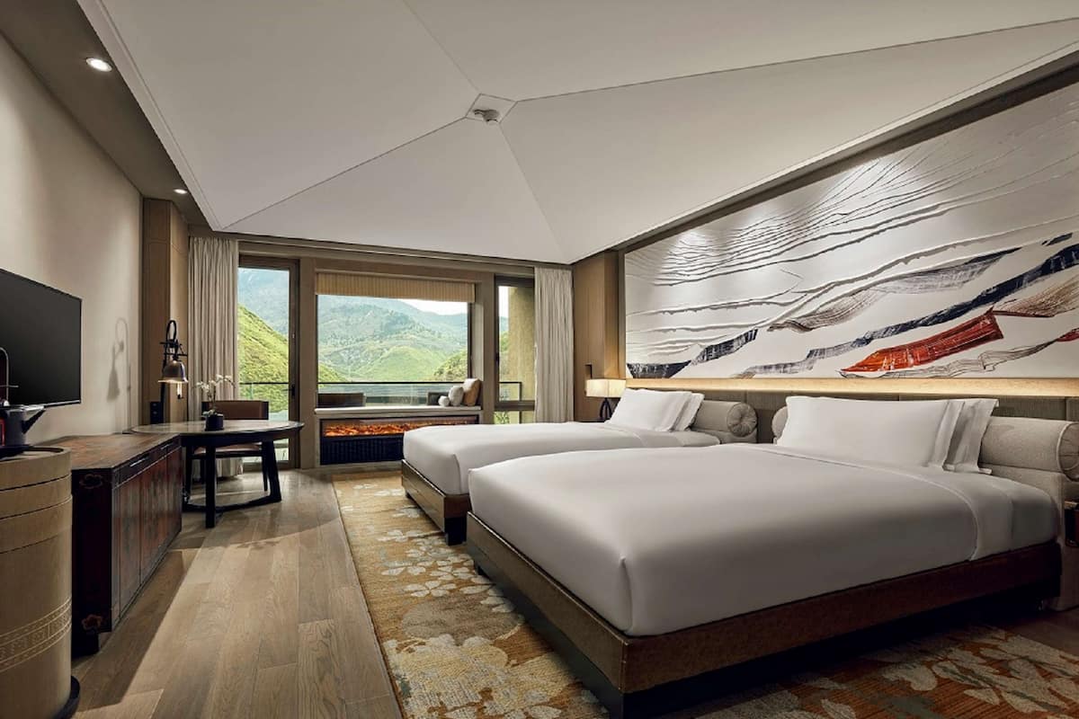 A hotel room with two beds, a desk, a TV, and a large window with a mountain view.