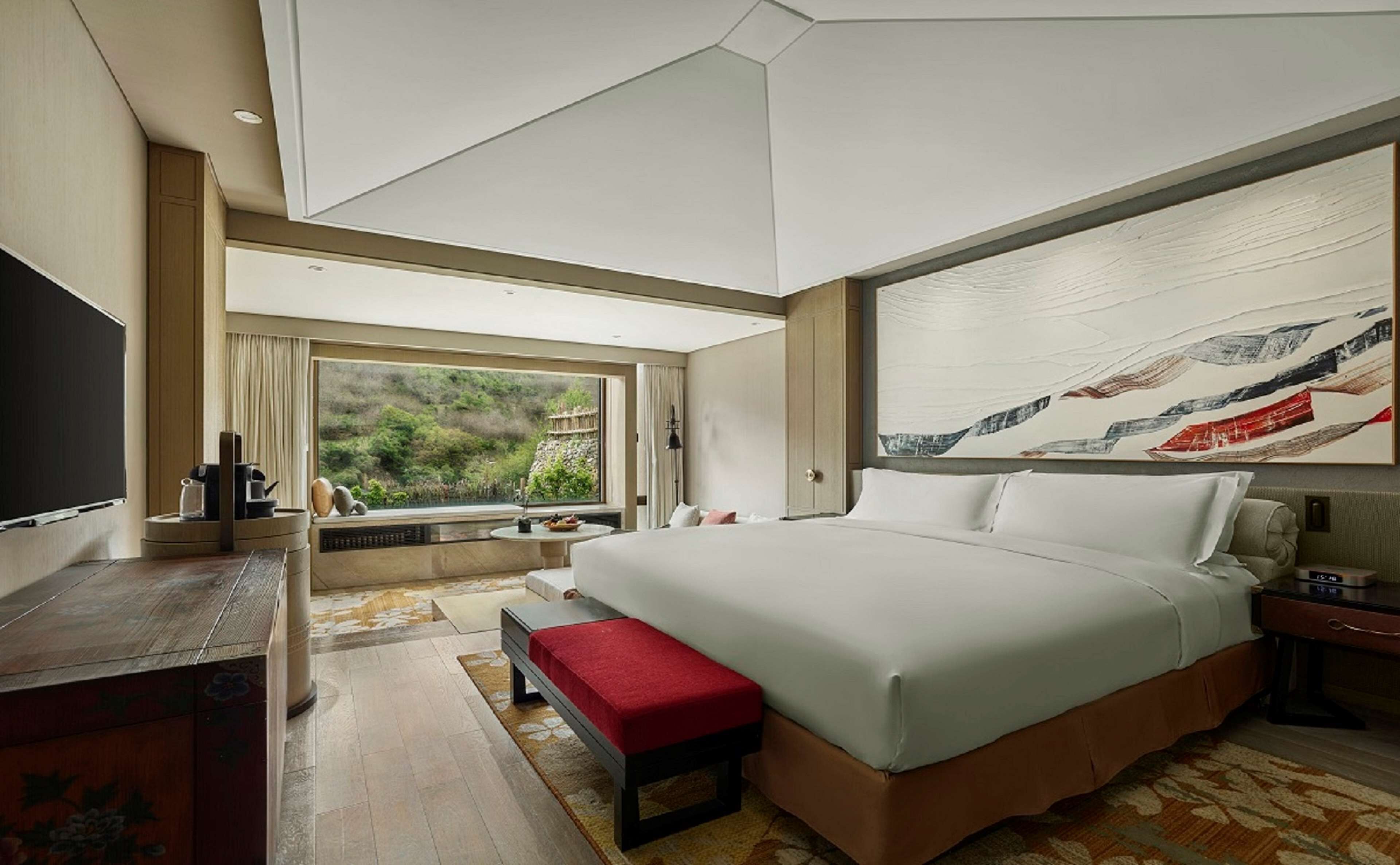 A modern hotel room with a large bed, a flat-screen TV, a dining area with a view, and a large abstract painting.
