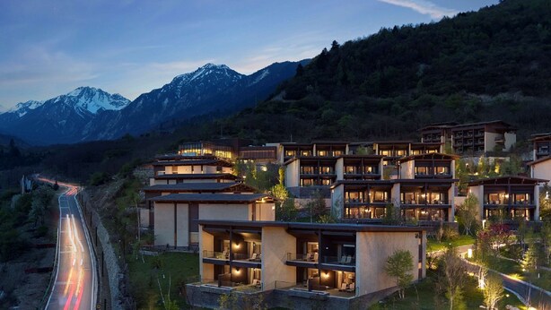 A modern hotel complex with multiple buildings, each featuring balconies and large windows, set against a backdrop of mountains and a winding road. - Conrad Jiuzhaigou (Ngawa Autonomous Prefecture)
